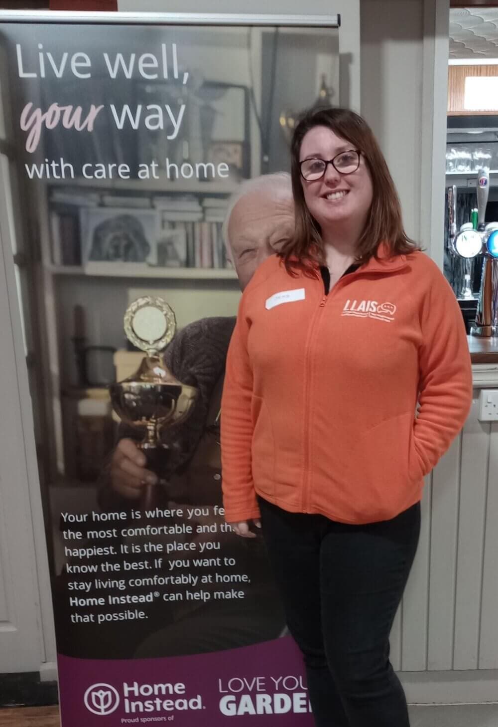 Young woman smiling in an orange jacket stands beside a "Home Instead" banner about home care services. - Home Instead