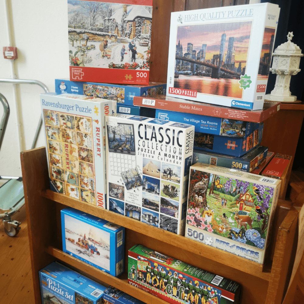 A shelf filled with various jigsaw puzzles, featuring a variety of artwork and box sizes, mostly 500 to 1500 pieces. - Home Instead