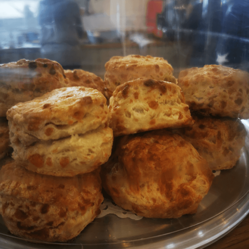 A stack of golden-brown scones displayed under a glass dome on a metal tray. - Home Instead