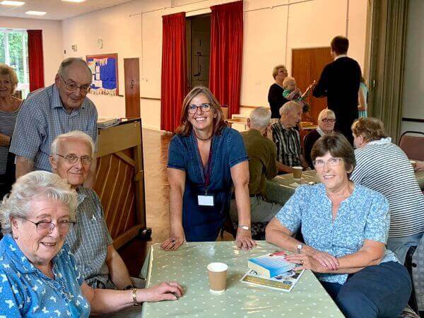 A group of elderly individuals and a middle-aged woman smile while sitting and standing around a table in a community hall. - Home Instead