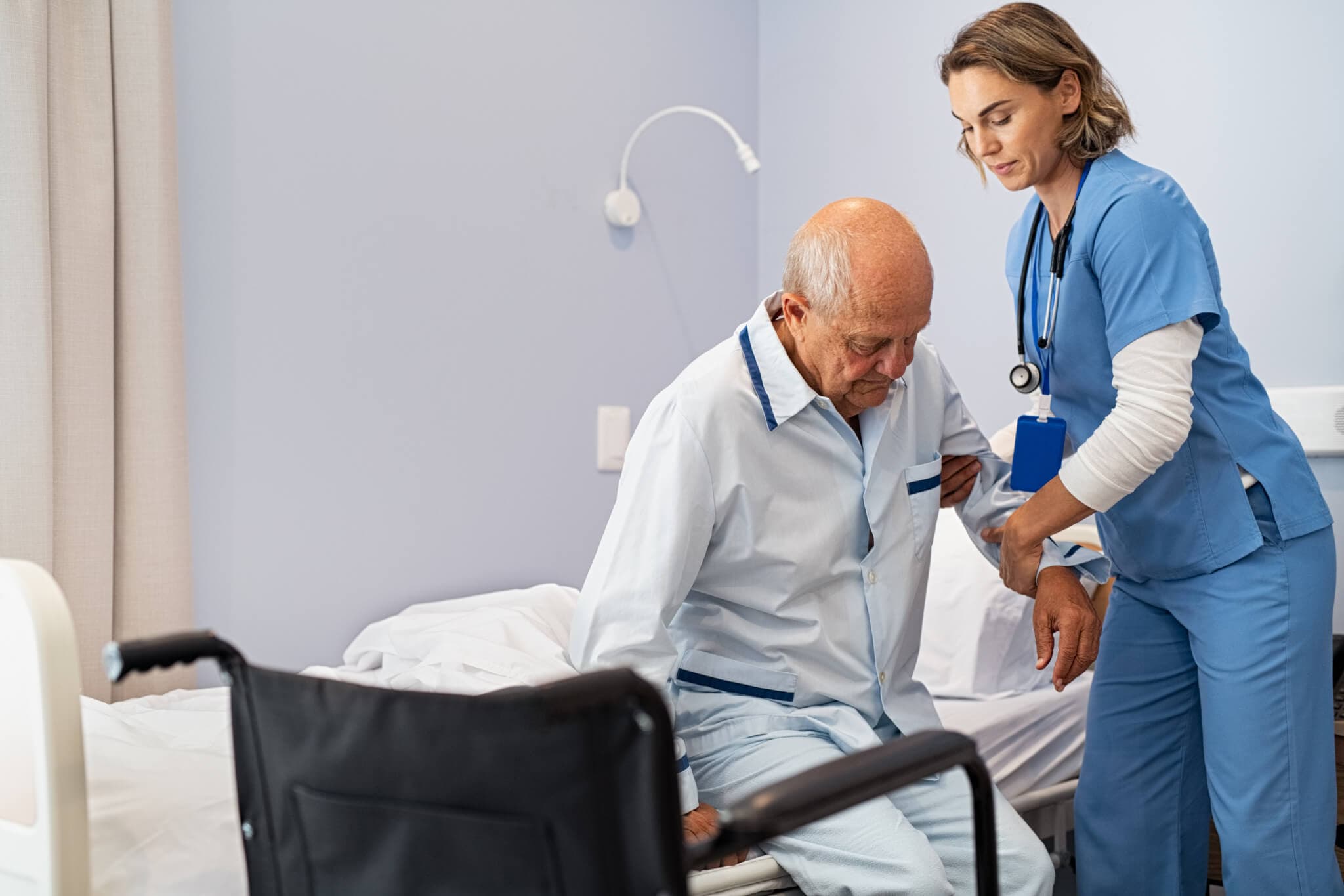 A nurse assists an elderly man as he gets up from a bed in a hospital room, with a wheelchair nearby. - Home Instead