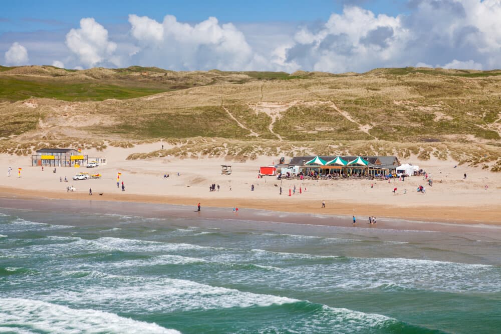 People enjoying a sunny day at a beach with a restaurant and colorful tents in the background, dunes and hills beyond. - Home Instead