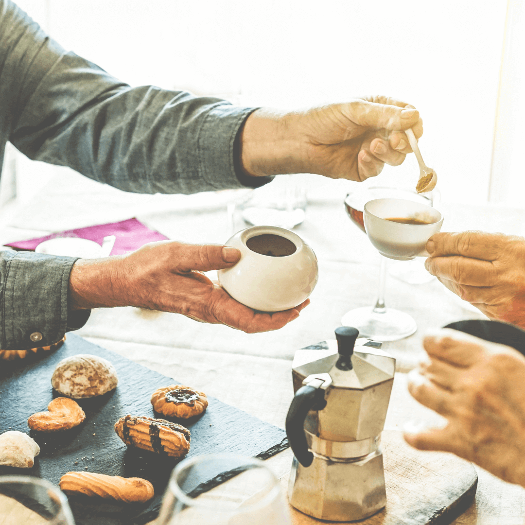 People enjoying coffee with assorted pastries on the table, one person adding sugar to their cup. - Home Instead