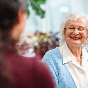 Elderly woman with white hair and glasses smiling, wearing a light blue cardigan, facing a blurred figure. - Home Instead