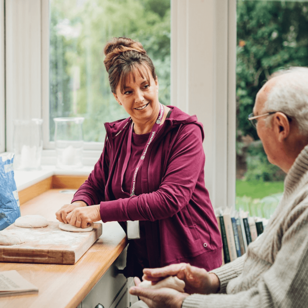 A woman in a purple jacket kneads dough on a kitchen counter, talking to an elderly man seated nearby. - Home Instead
