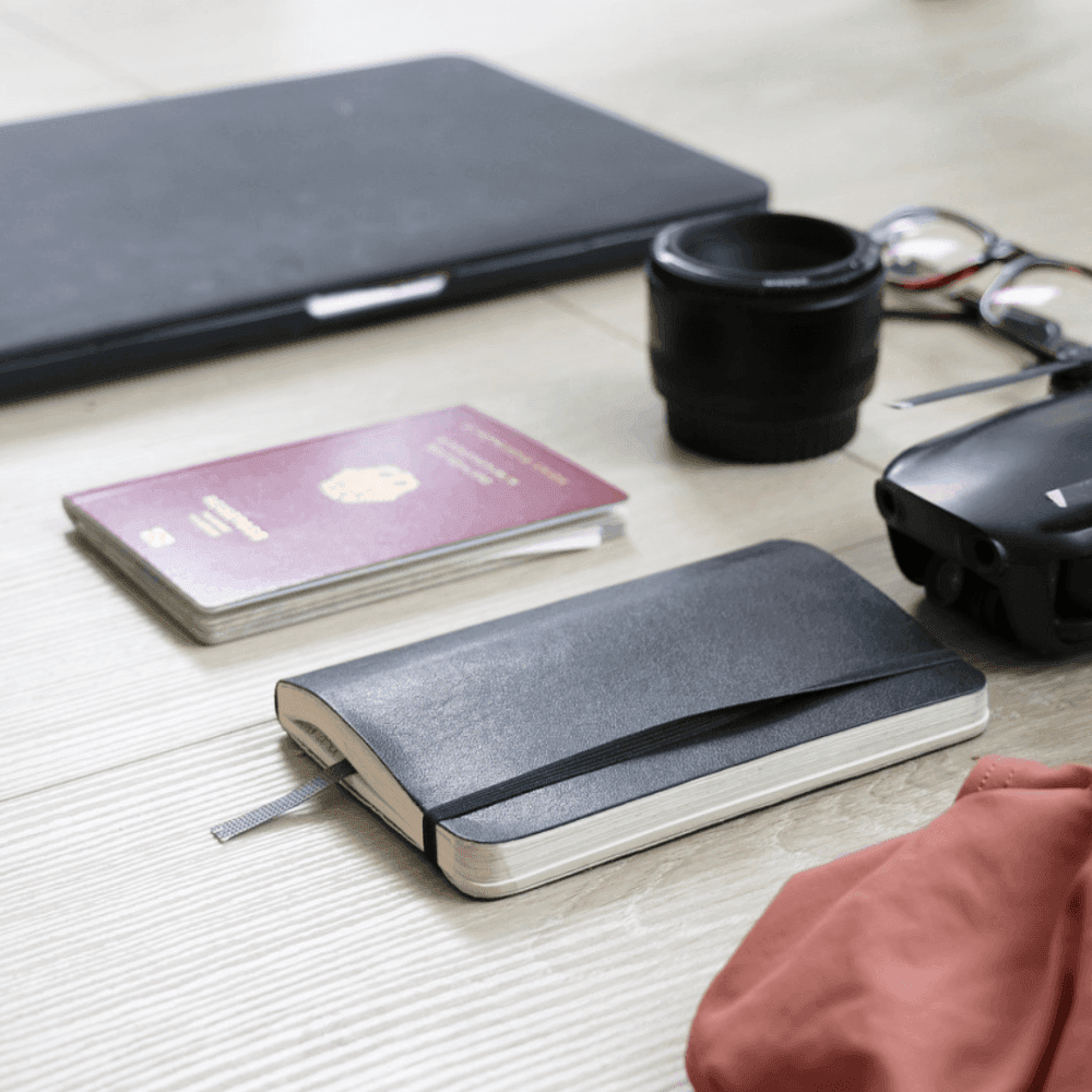 Travel essentials including a notebook, passport, camera lens, and sunglasses arranged on a light wooden surface. - Home Instead