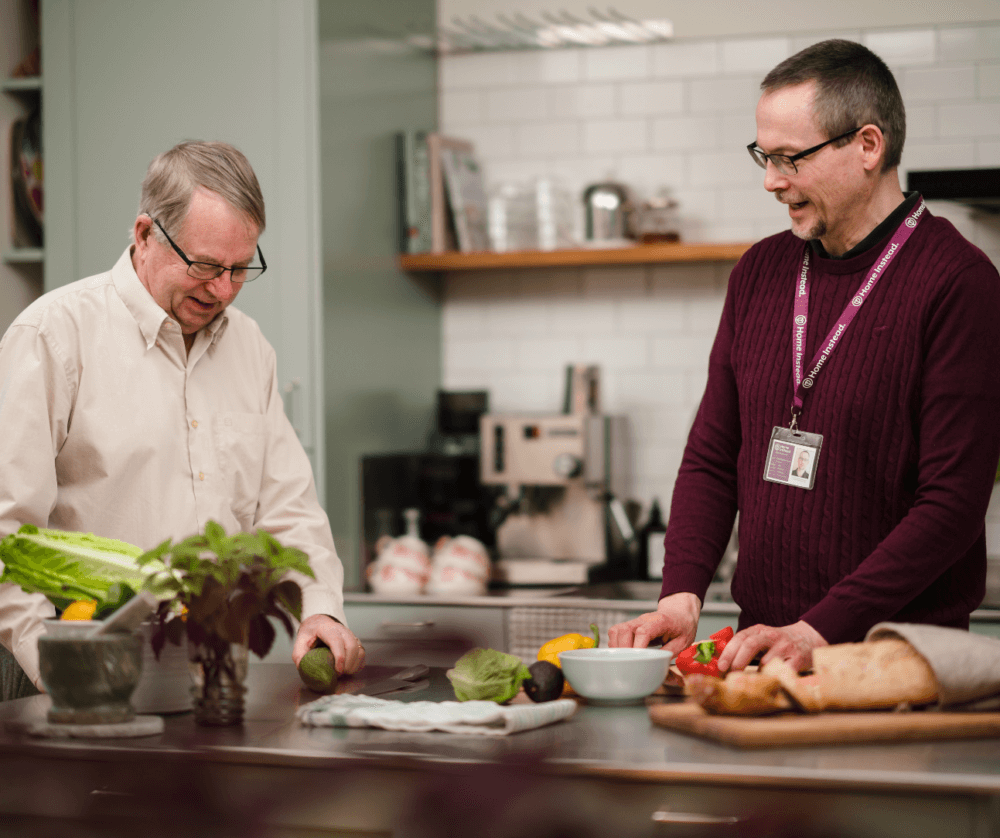 Two men are preparing food in a modern kitchen, talking and smiling, with various ingredients on the counter. - Home Instead