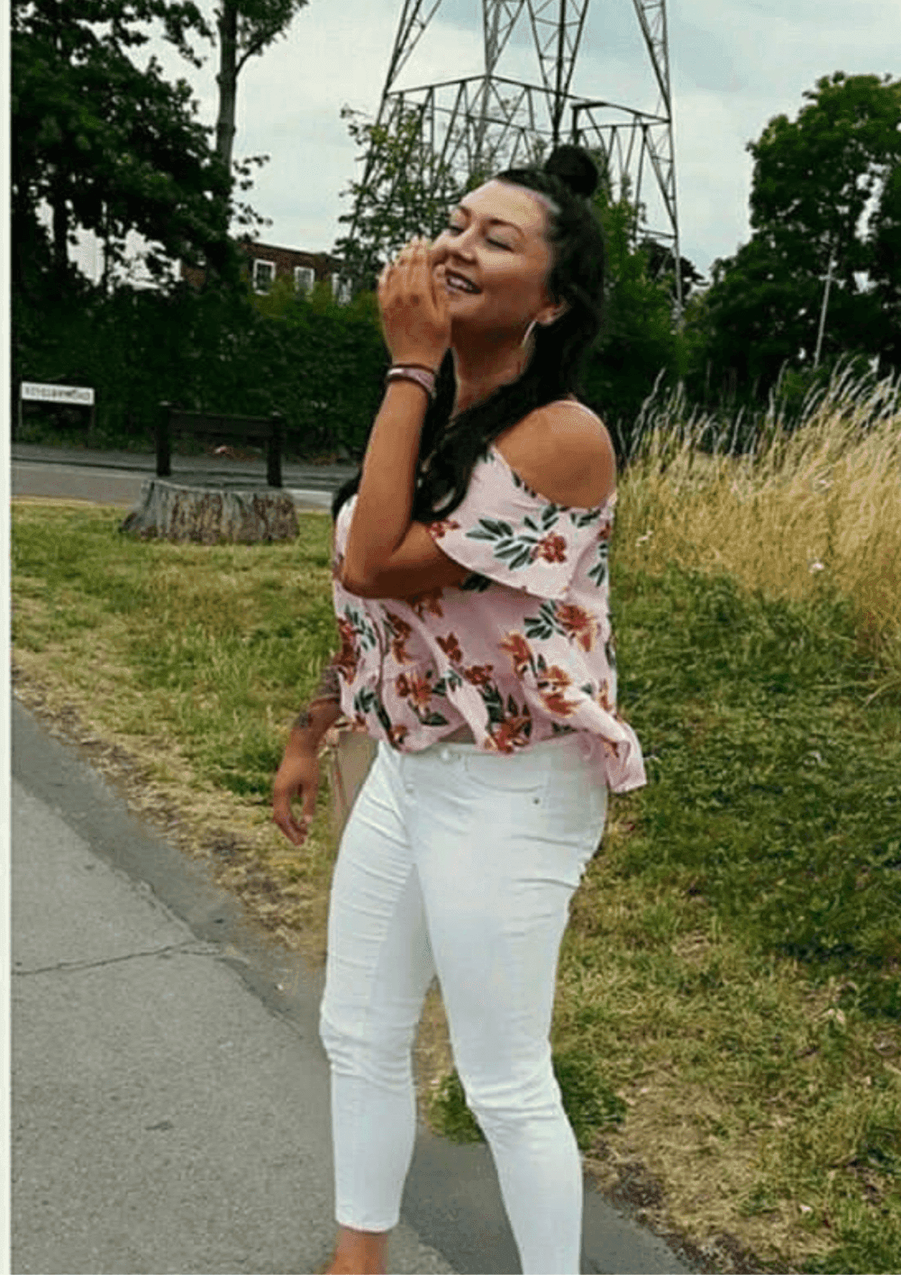 A woman in a floral off-shoulder top and white pants stands outdoors, smiling and laughing, with a grassy area behind her. - Home Instead