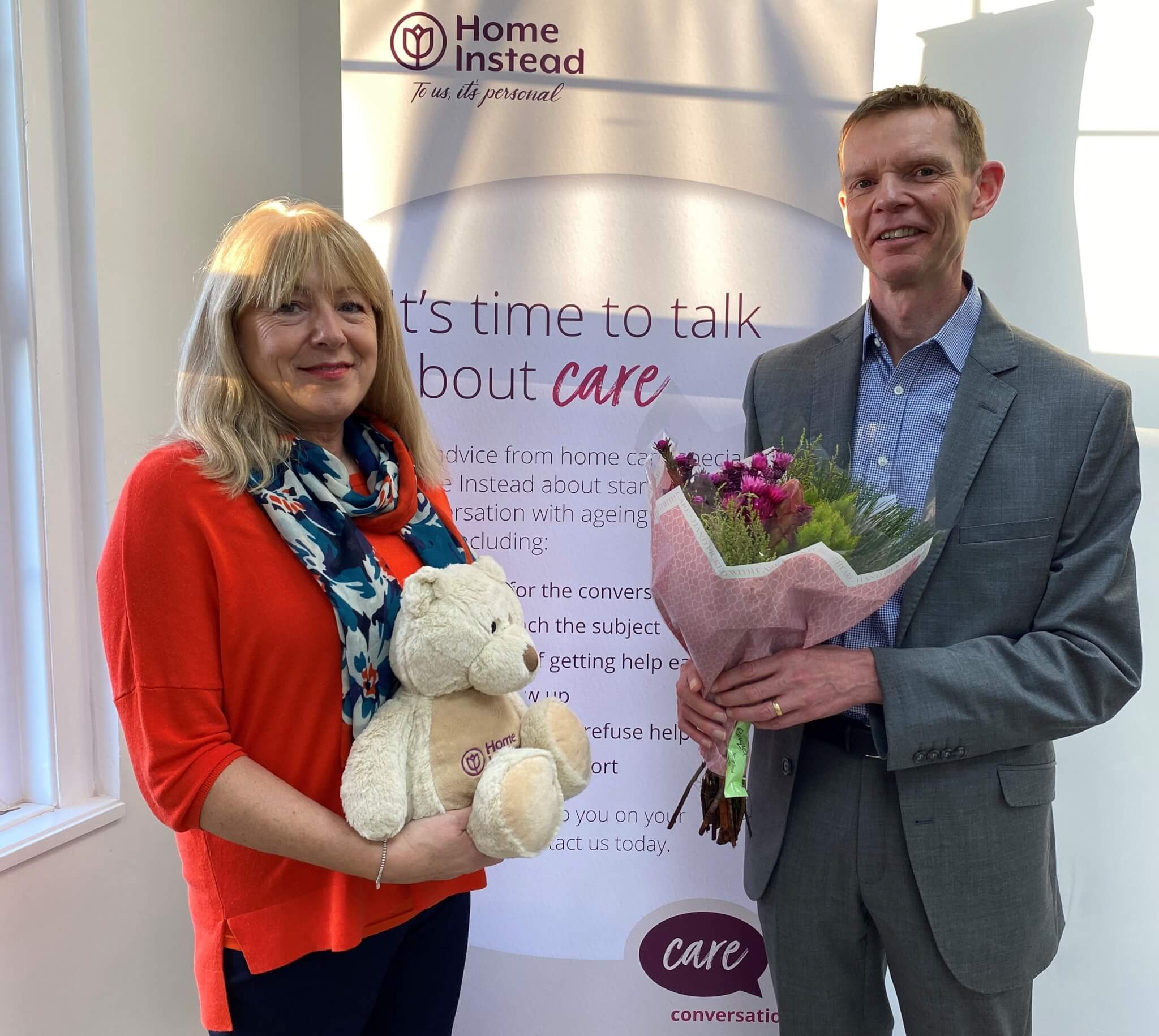 Two people standing in front of a "Home Instead" banner, holding a bouquet and a teddy bear. - Home Instead