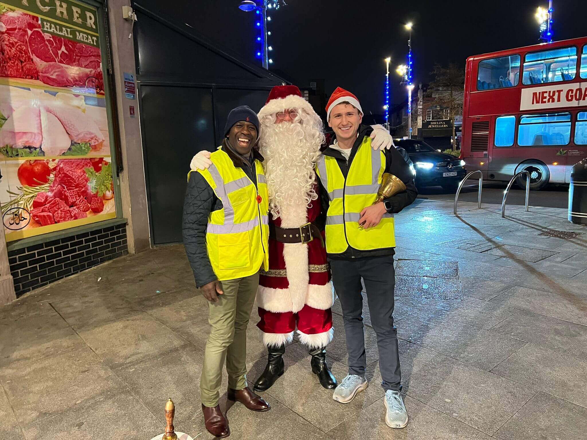 Two men in yellow vests stand with a person dressed as Santa Claus on a city street at night. - Home Instead