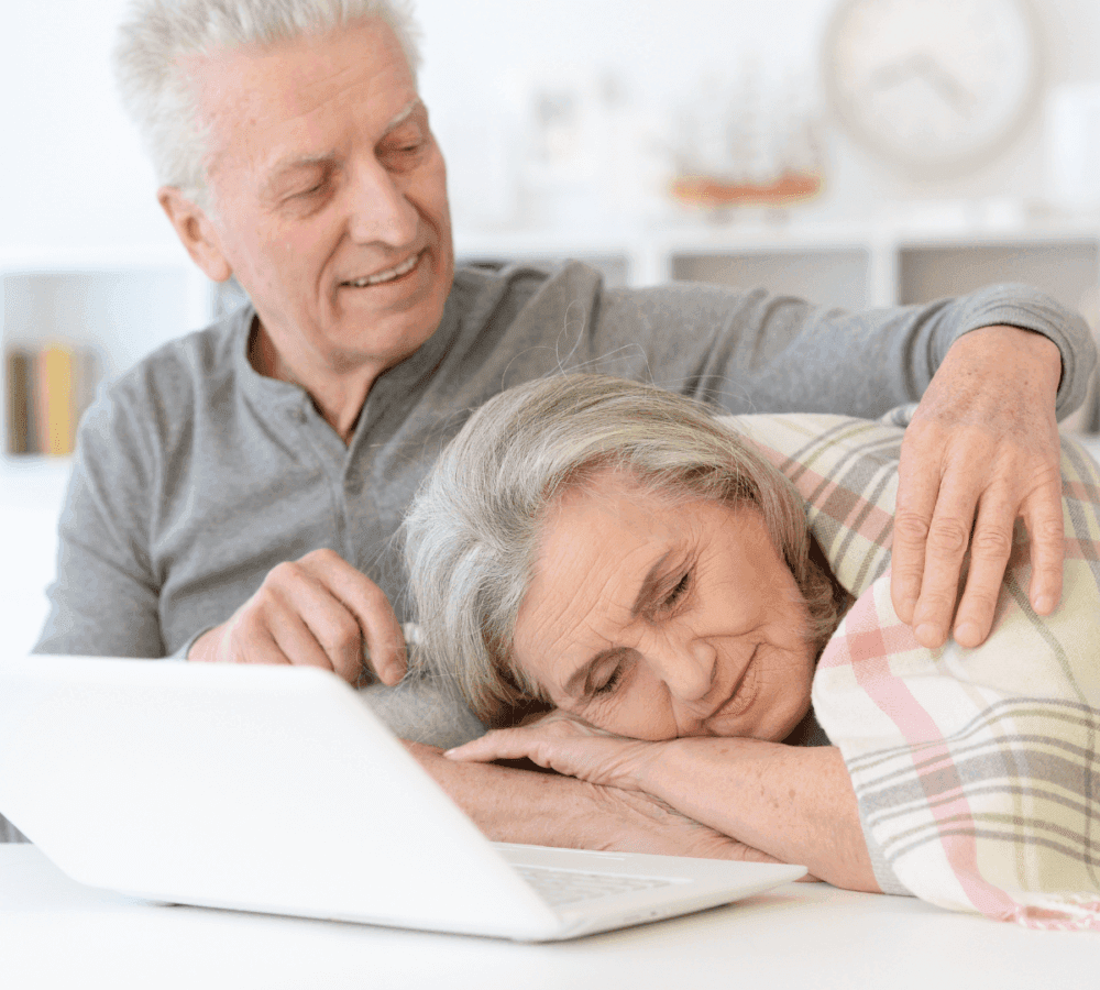 An elderly man affectionately holds an elderly woman who is resting her head on her arms near an open laptop. - Home Instead
