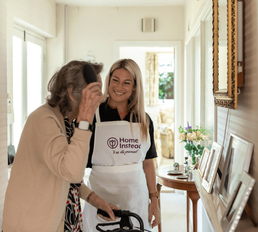 A caregiver in a "Home Instead" uniform assists an elderly woman with a walker in a warmly-lit hallway. - Home Instead