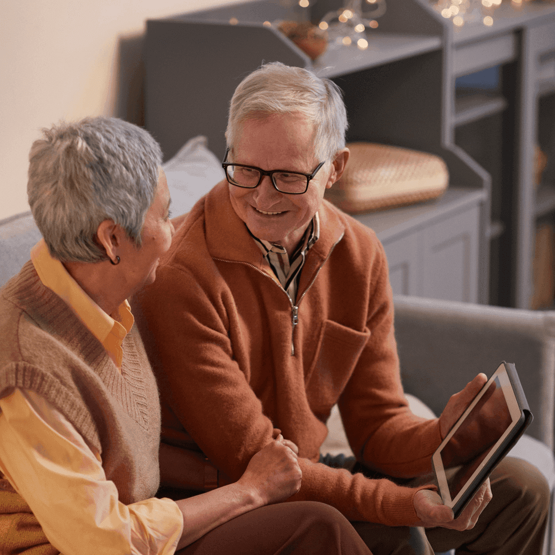 An elderly couple sits on a couch, smiling and holding a tablet in their cozy living room. - Home Instead