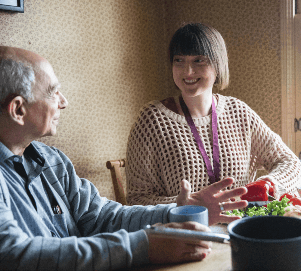 Two people sitting at a table talking, with vegetables and a cup on the table in front of them. - Home Instead