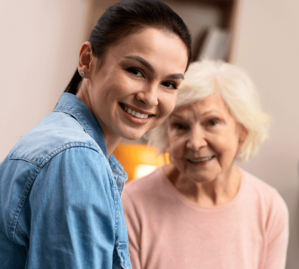 Younger woman in denim shirt smiling, sitting beside an older woman wearing a pink top, both looking at the camera. - Home Instead