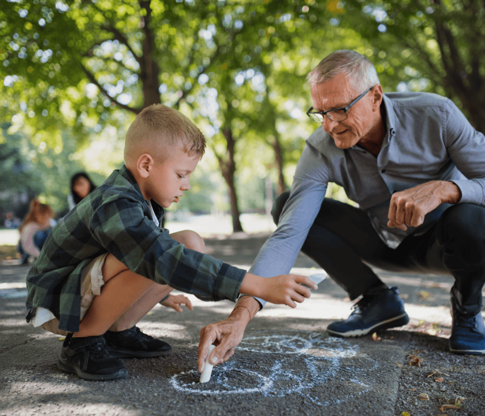 An elderly man and a young boy drawing with chalk on a sidewalk in a park with trees in the background. - Home Instead