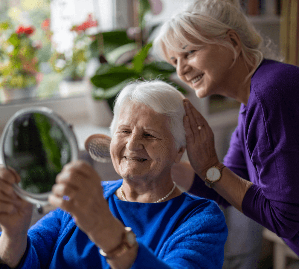 Two elderly women smiling, with one holding a mirror while the other gently touches her shoulder in a brightly lit room. - Home Instead