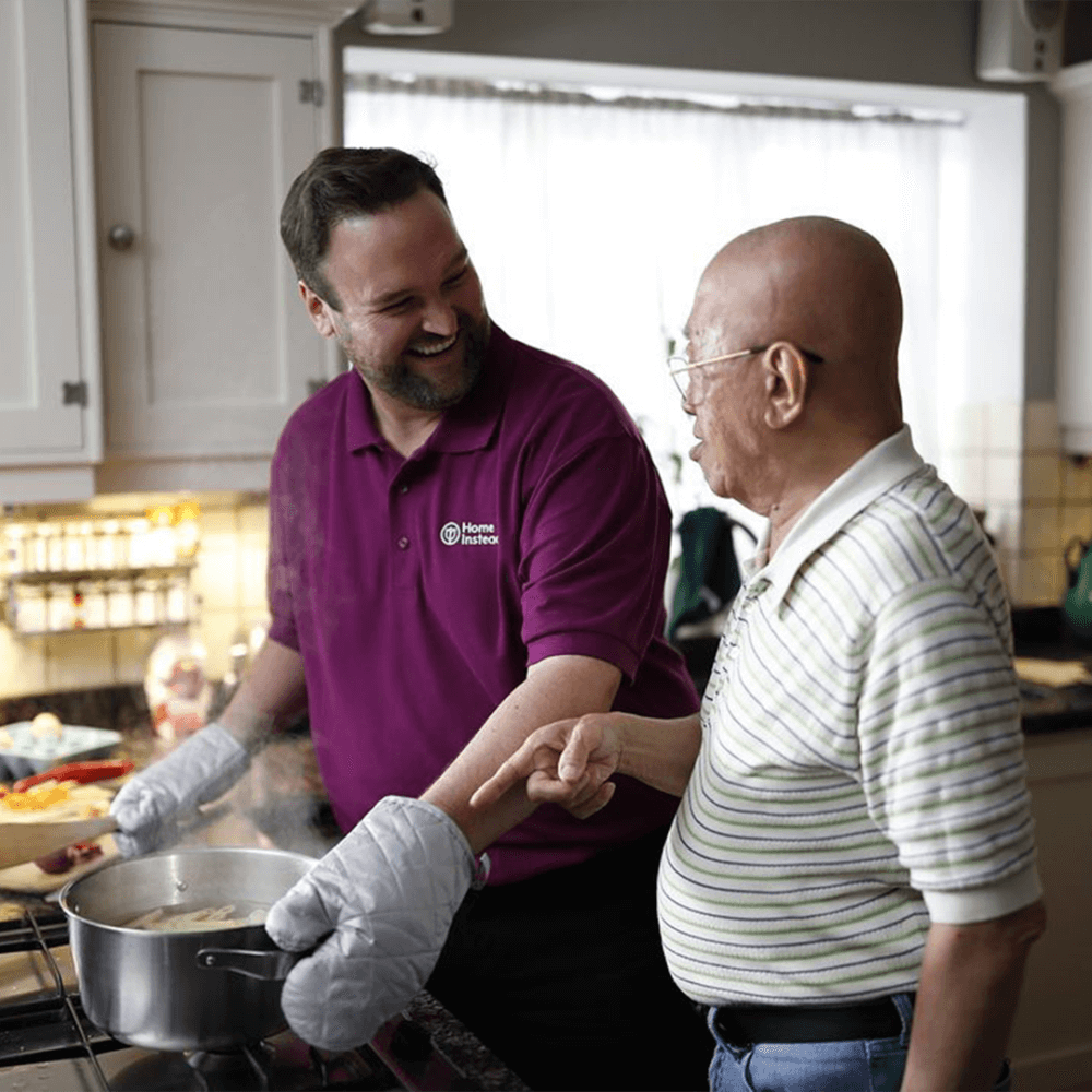 A caregiver in a purple shirt assists an elderly man with cooking in a kitchen, smiling and engaged in conversation. - Home Instead