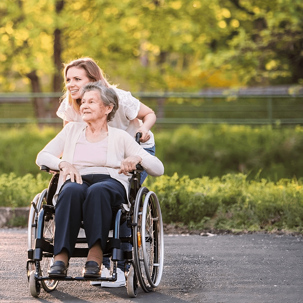 A young woman joyfully pushes an elderly woman in a wheelchair outdoors on a sunny day with green trees in the background. - Home Instead