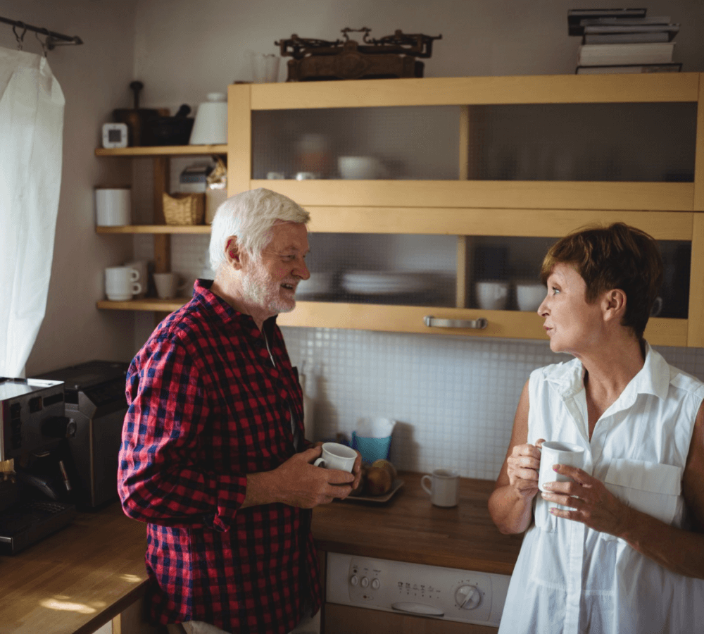 Two people drinking coffee and talking in a kitchen with wooden cabinets and a coffee maker. - Home Instead