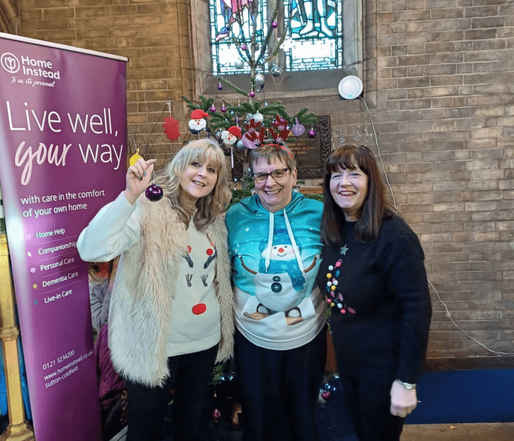 Three smiling women in festive sweaters stand in front of a decorated Christmas tree and a "Live well, your way" banner. - Home Instead