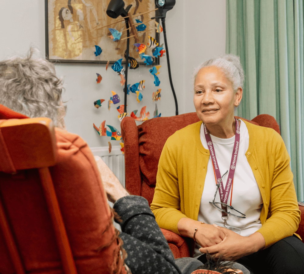 A caregiver in a yellow cardigan talks with an elderly person sitting in a red chair in front of her. - Home Instead