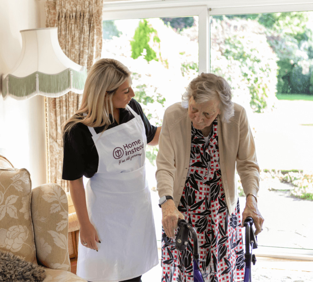 A caregiver assists an elderly woman with a walker in a well-lit living room with large windows and garden view. - Home Instead
