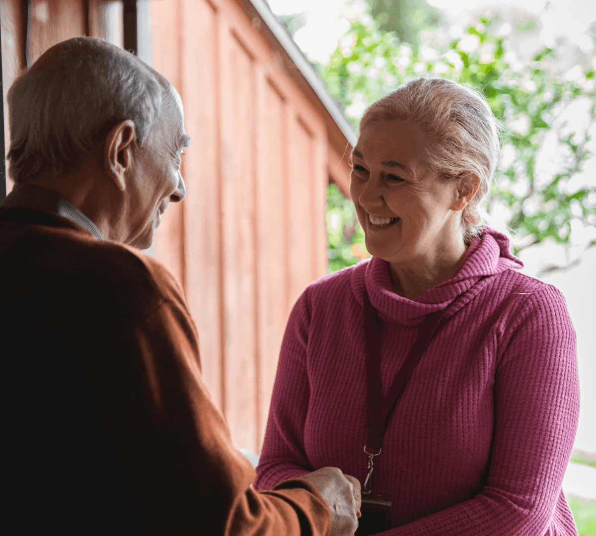 Two people, one in a pink sweater and the other in a brown sweater, smiling and shaking hands outdoors. - Home Instead