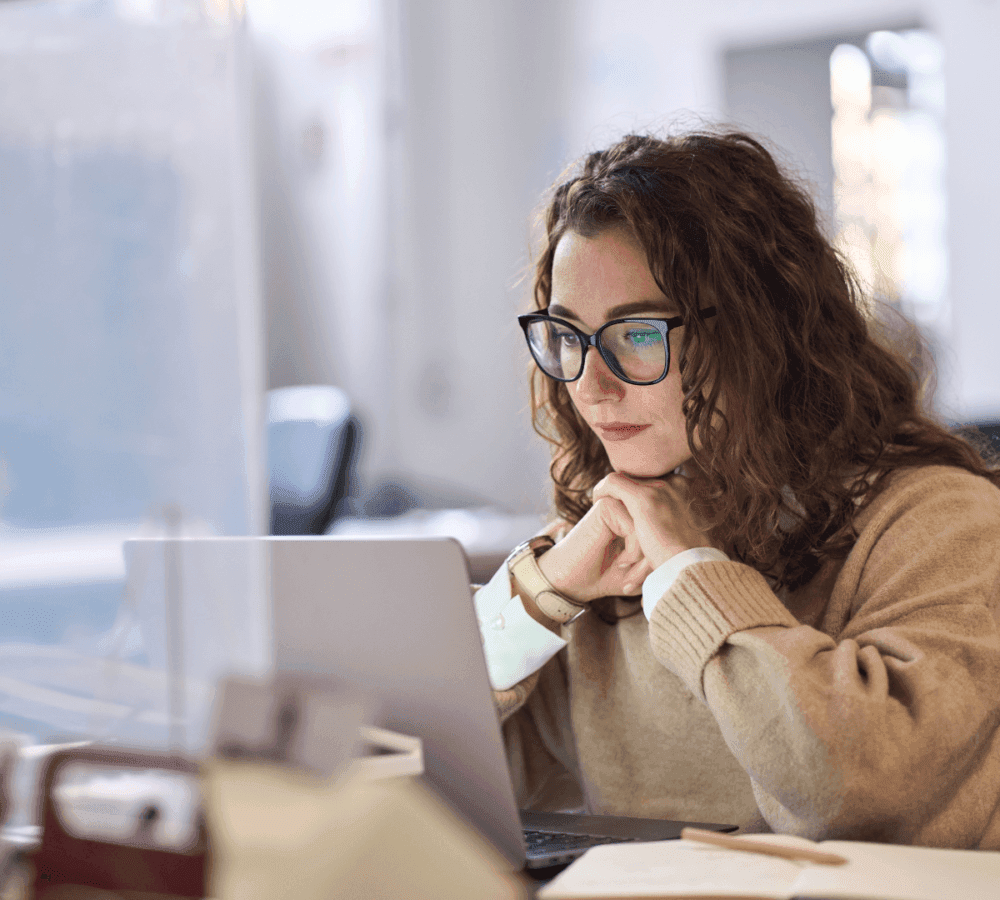 A woman with curly hair and glasses works intently on a laptop at a desk. - Home Instead