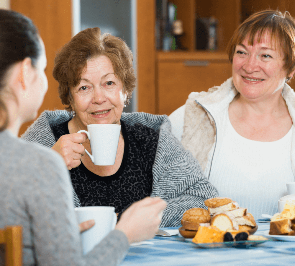 Three women enjoying coffee and pastries at a table, smiling and engaged in conversation. - Home Instead