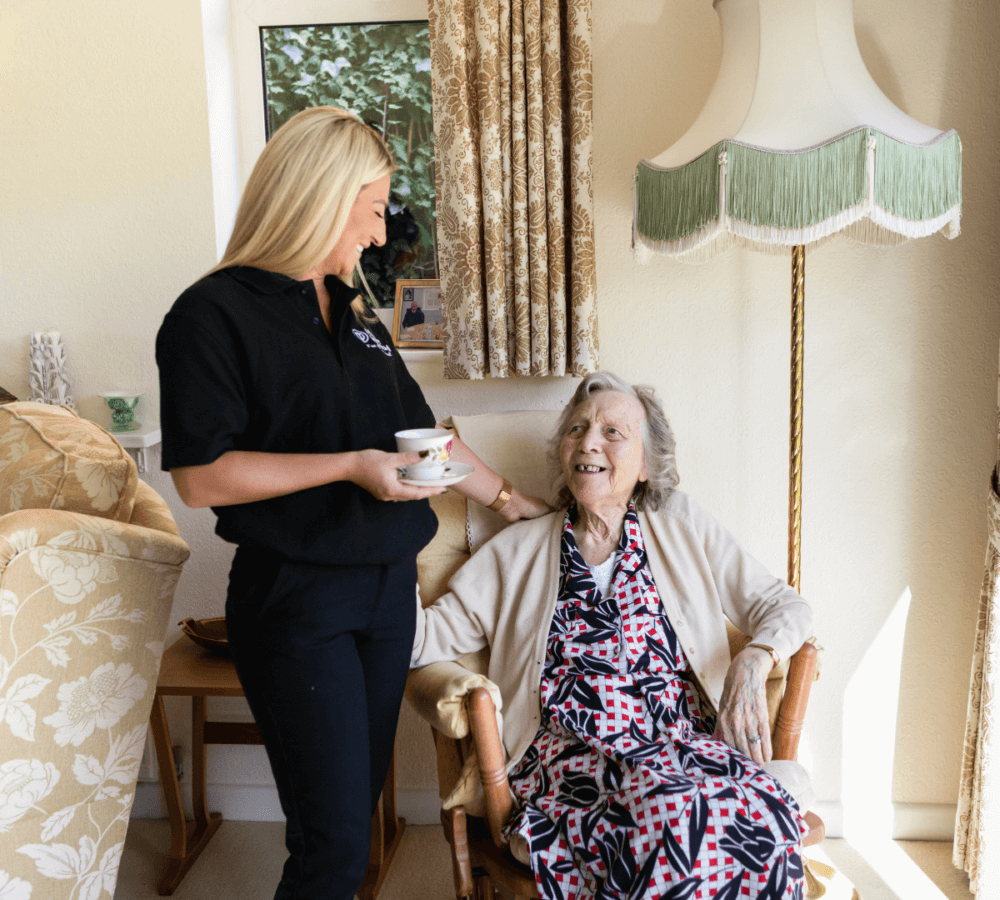 A young woman smiling and holding a cup, standing next to an elderly woman seated in a cozy living room. - Home Instead