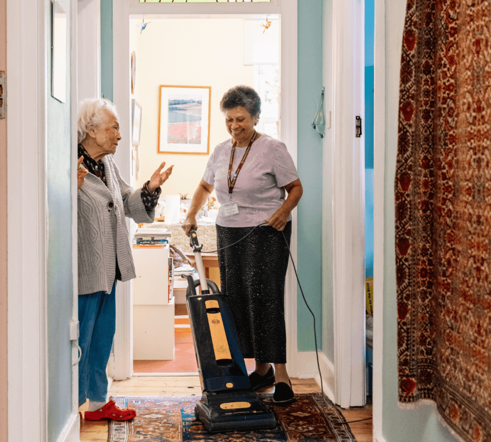 An older woman talks enthusiastically with a housekeeper vacuuming a hallway in a brightly lit home. - Home Instead