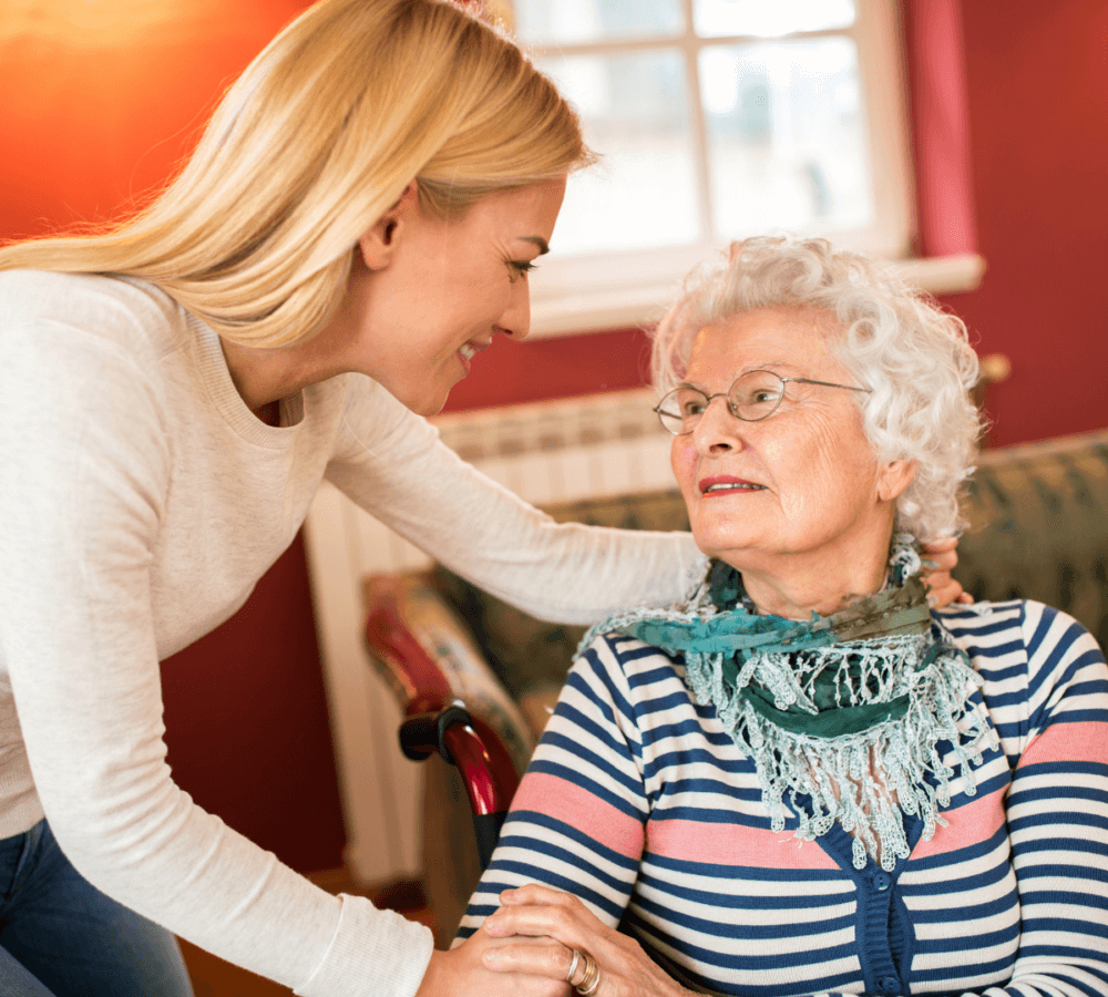 A young woman smiles and leans towards an elderly woman seated in a wheelchair, both sharing a warm moment indoors. - Home Instead