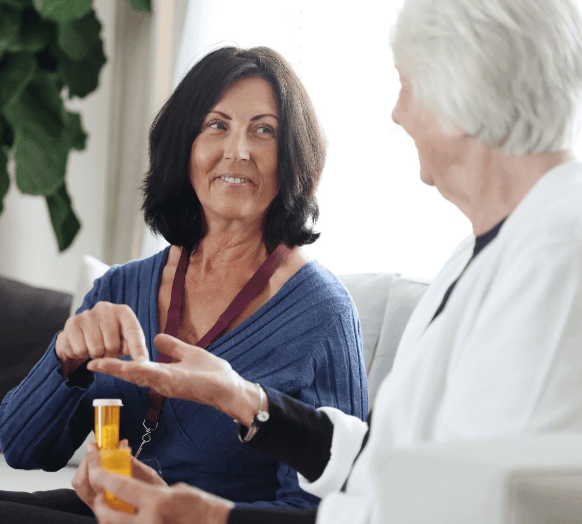 A woman helps an older adult with medication while they sit on a couch, both smiling at each other. - Home Instead