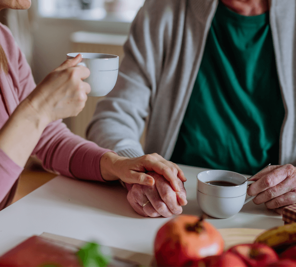 Two people holding hands and drinking coffee at a table with fruit. Only their hands and torsos are visible. - Home Instead