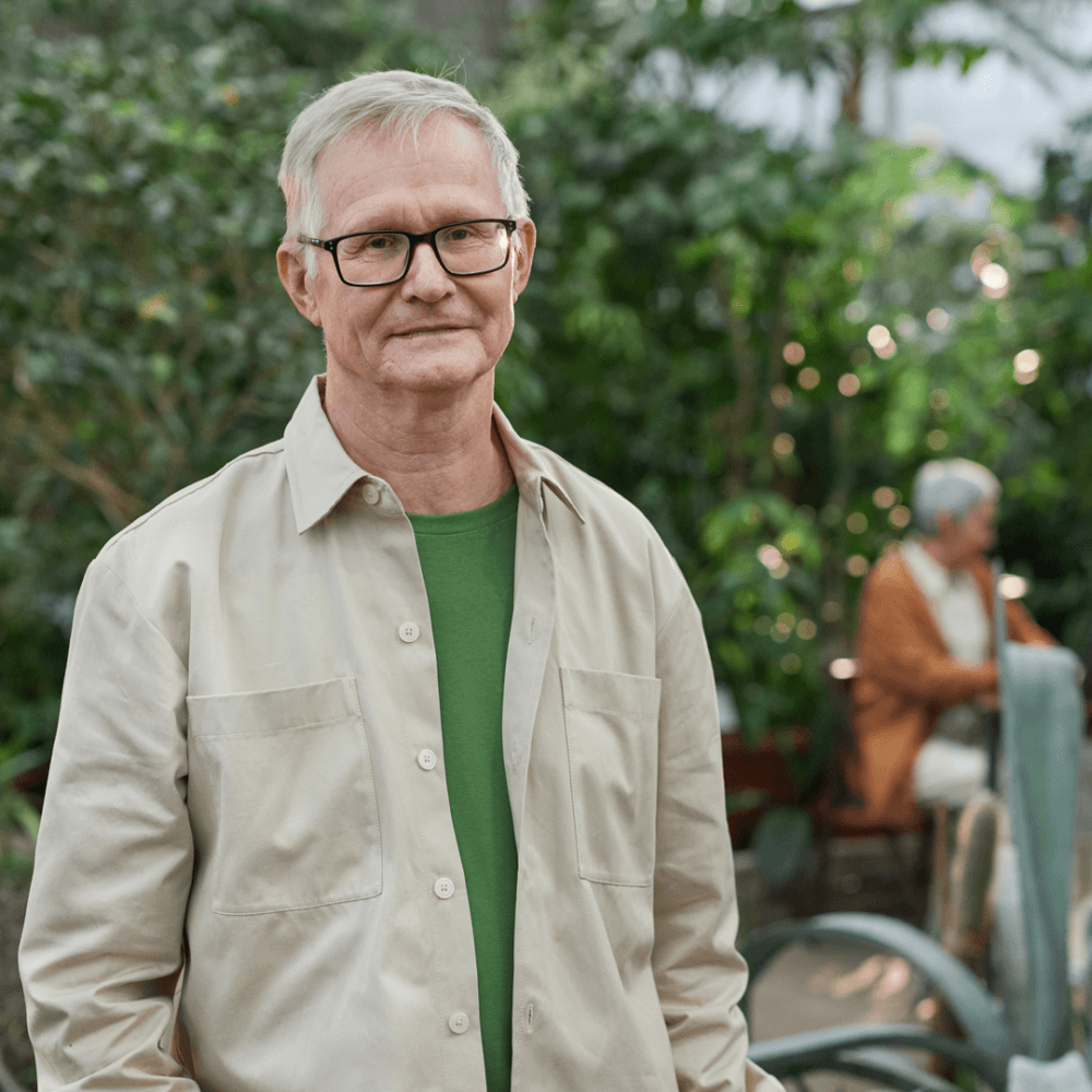 An elderly man with glasses and a beige shirt stands outdoors in a garden with greenery. Another person sits in the background. - Home Instead