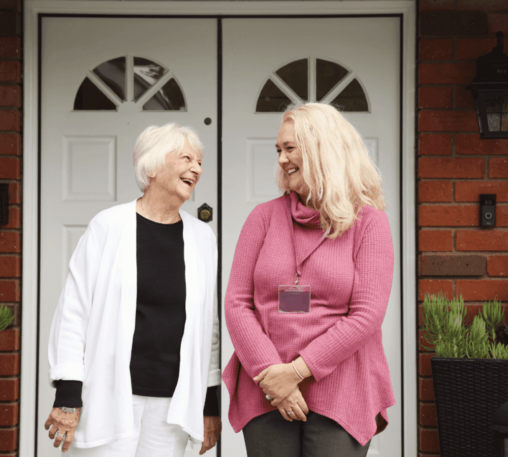 Two women, one older and one younger, laugh together in front of a house with a double white door. - Home Instead