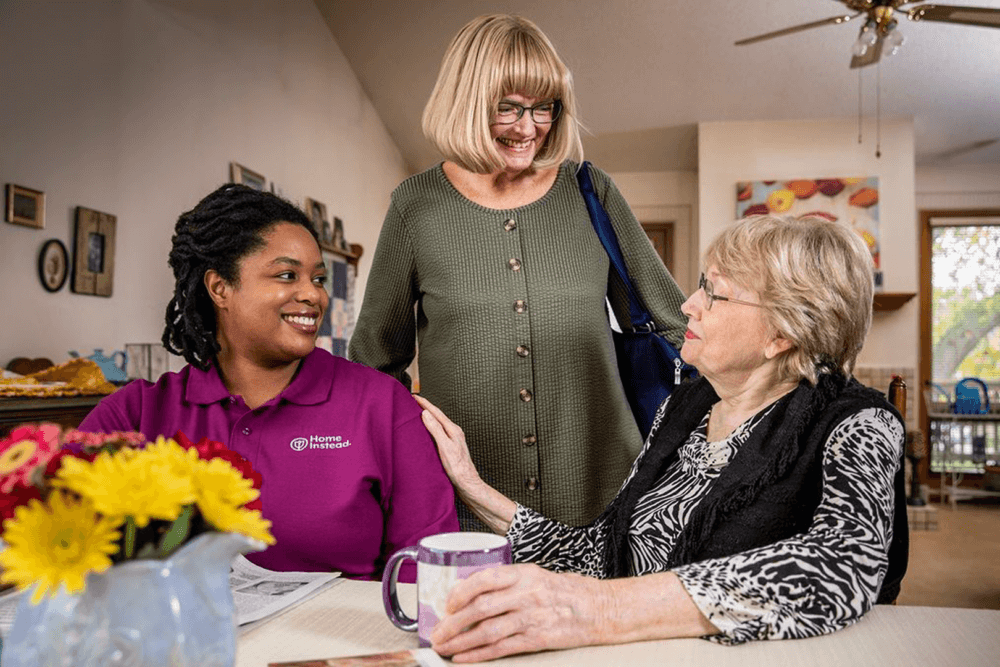 A caregiver in a purple shirt chats with two smiling elderly women in a cozy, well-lit room with a flower vase on a table. - Home Instead