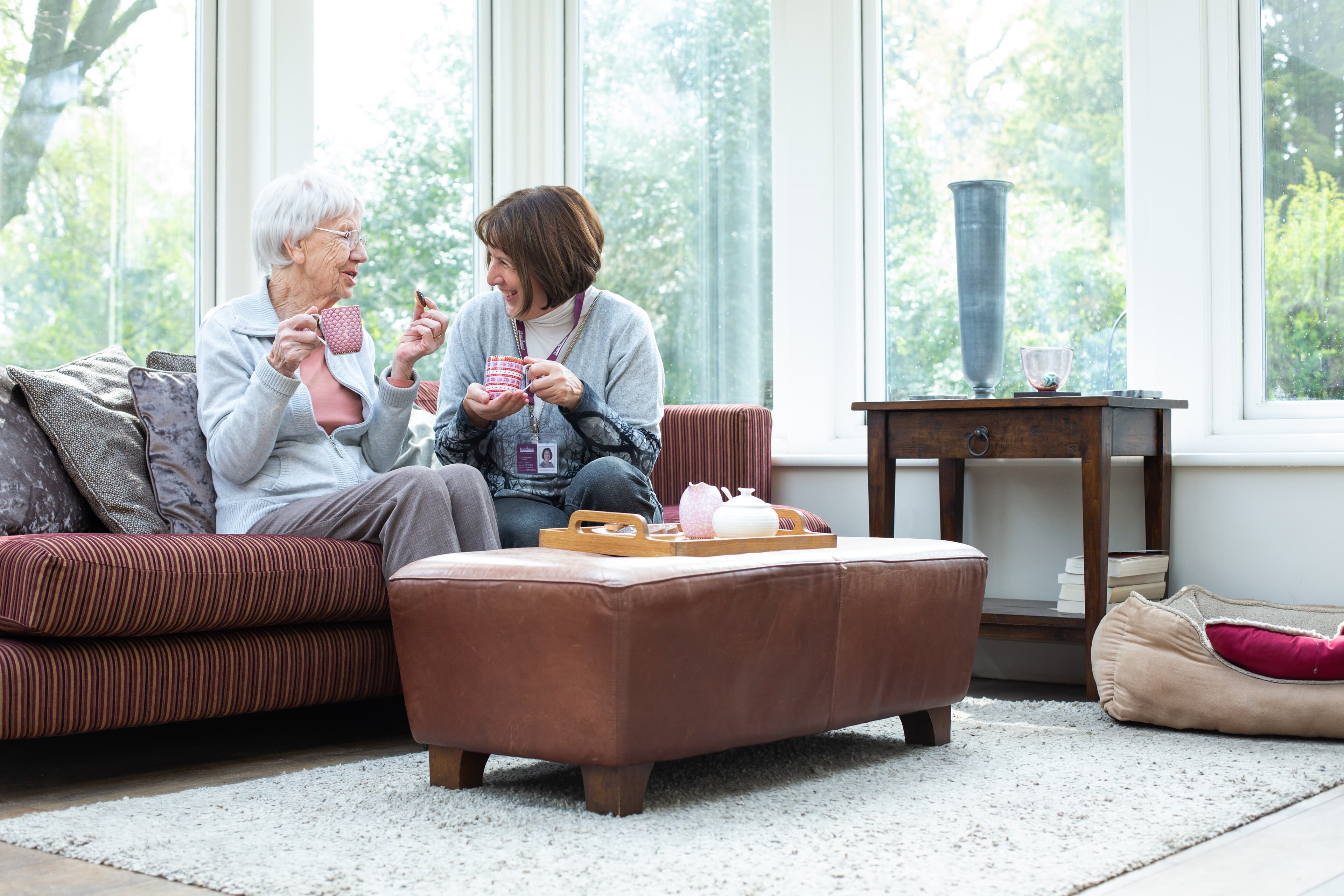 An elderly woman and a younger woman are smiling and sharing tea in a cozy, bright living room. - Home Instead