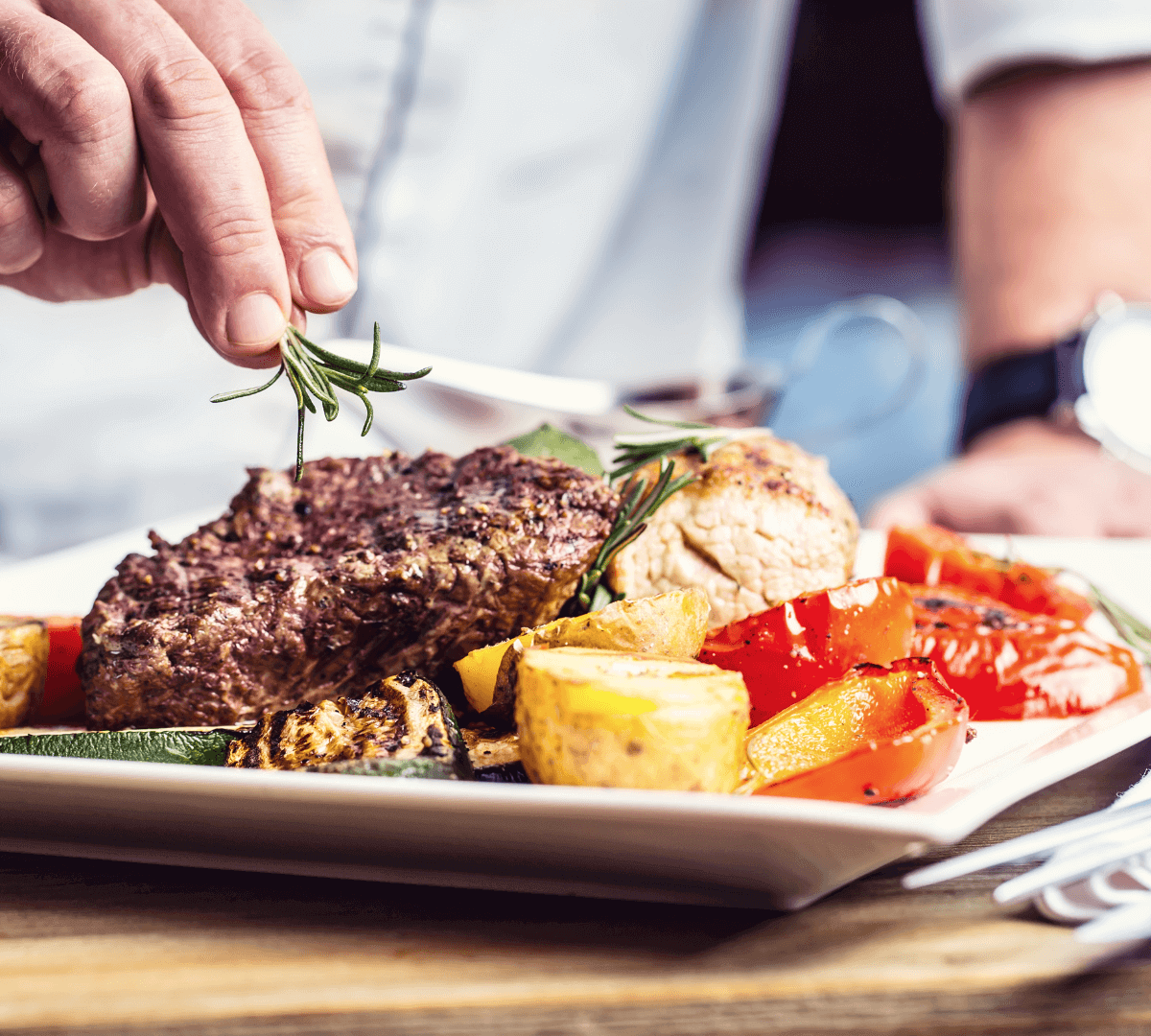 Chef garnishes grilled steak with rosemary on a plate of roasted vegetables, including potatoes, zucchini, and bell peppers. - Home Instead