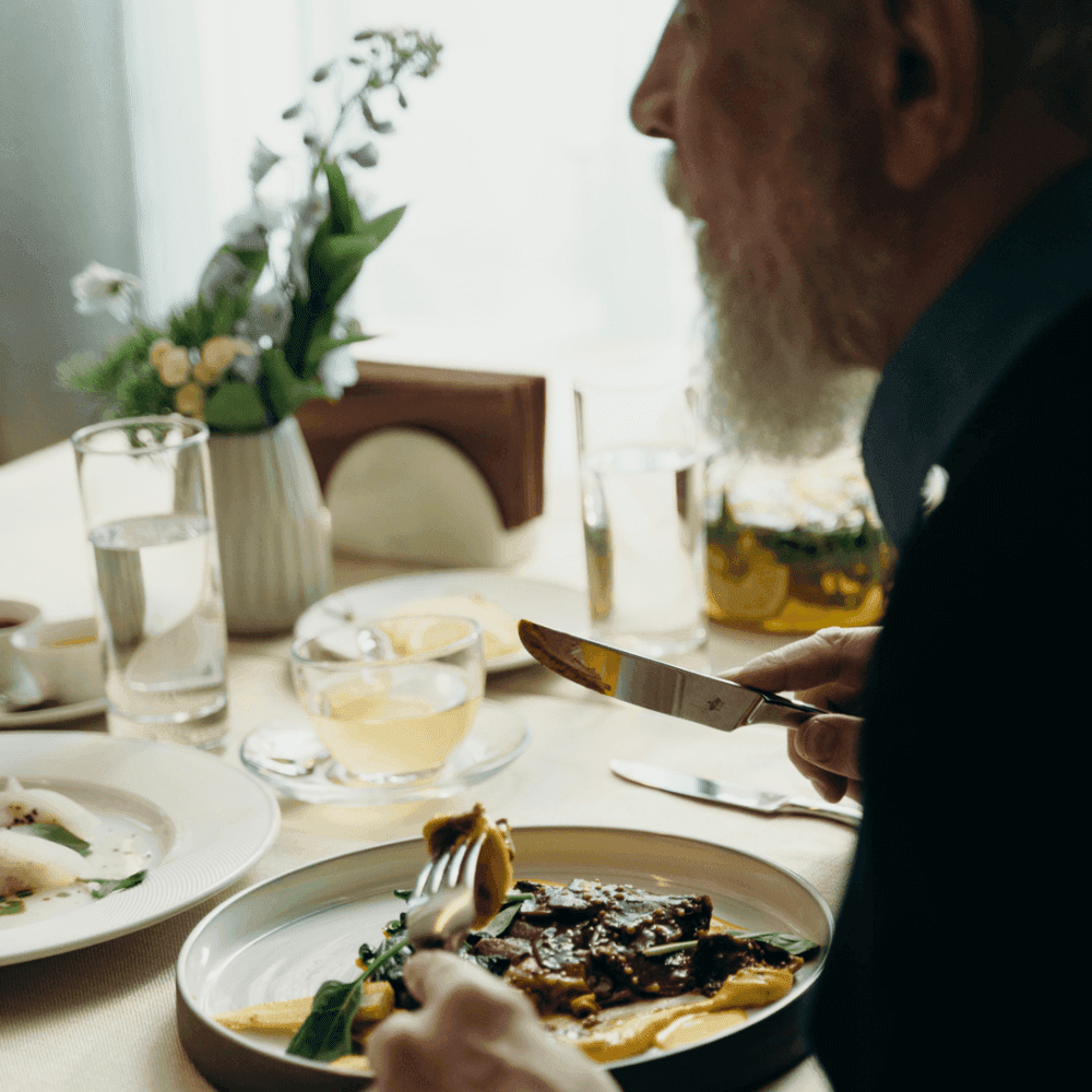A man with a beard dines at a table with plates of food, a glass of water, and a flower arrangement in the background. - Home Instead