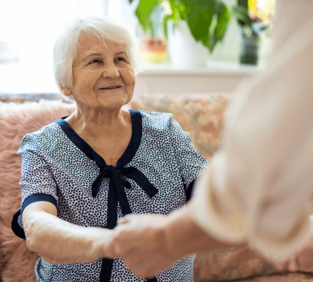 Elderly woman with short white hair smiling and holding the hand of another person indoors with plants in the background. - Home Instead