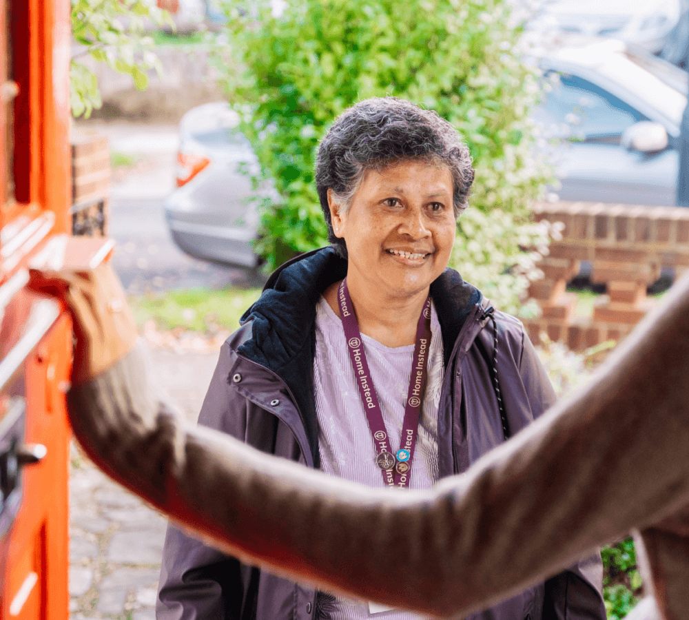 Person with short gray hair and lanyard smiling while standing outside an open door. - Home Instead