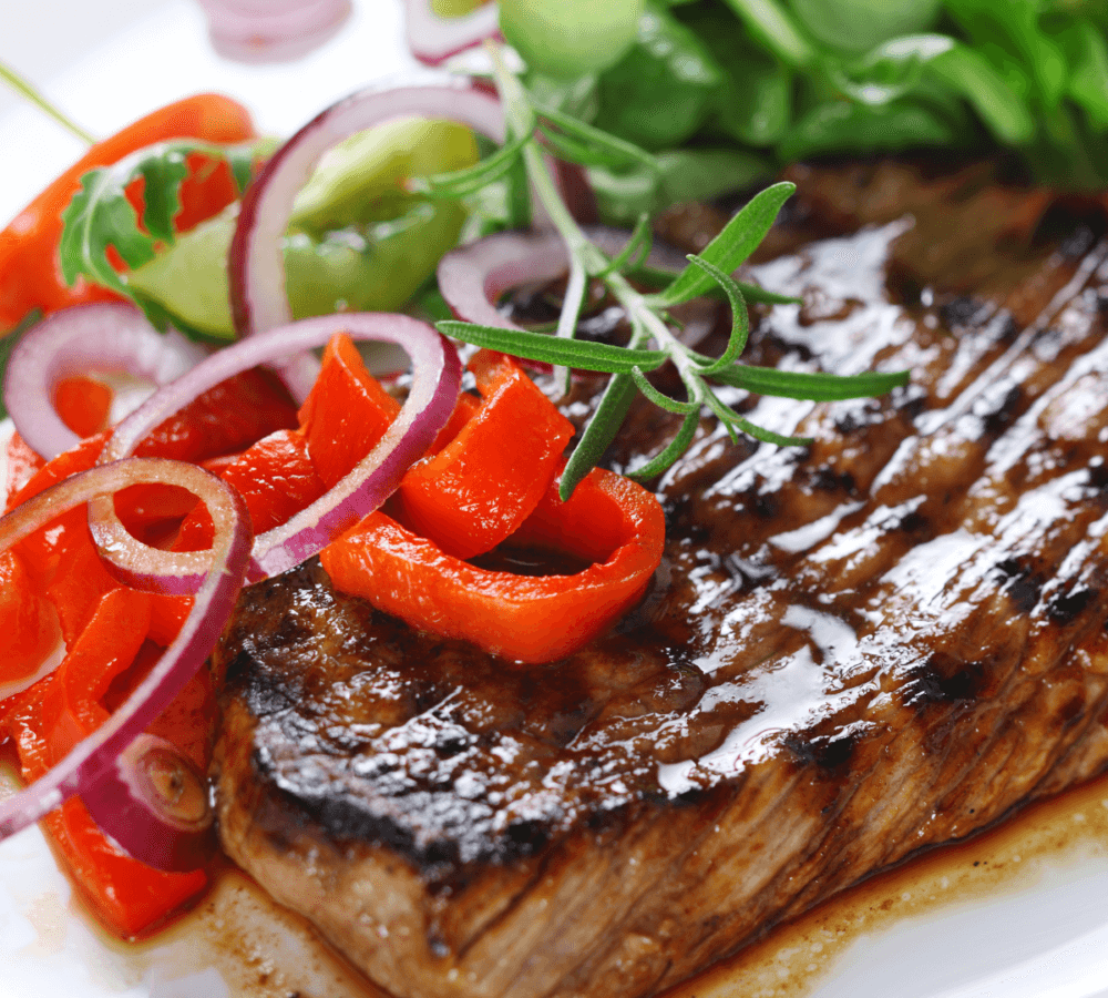 Grilled steak served with sliced red bell peppers, red onions, greens, and a sprig of rosemary on a white plate. - Home Instead