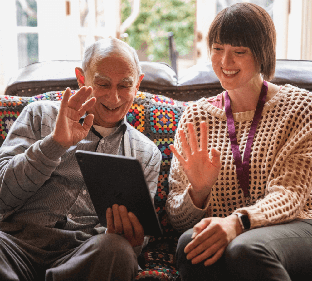 An elderly man and a woman smiling and waving at a tablet while sitting on a colorful crochet blanket. - Home Instead