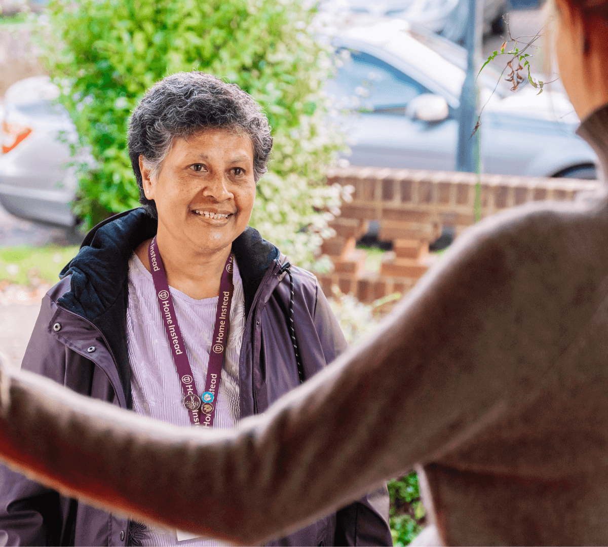 Smiling woman with short gray hair wearing a lanyard is greeted at a doorstep near a parked car and green bushes. - Home Instead