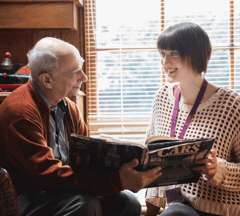 An elderly man and a young woman are smiling while looking at a magazine titled "CARS" by a sunlit window. - Home Instead