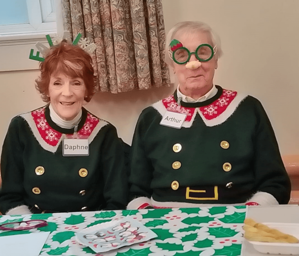 Two elderly people dressed in festive outfits, one wearing reindeer antlers and the other wearing novelty glasses. - Home Instead
