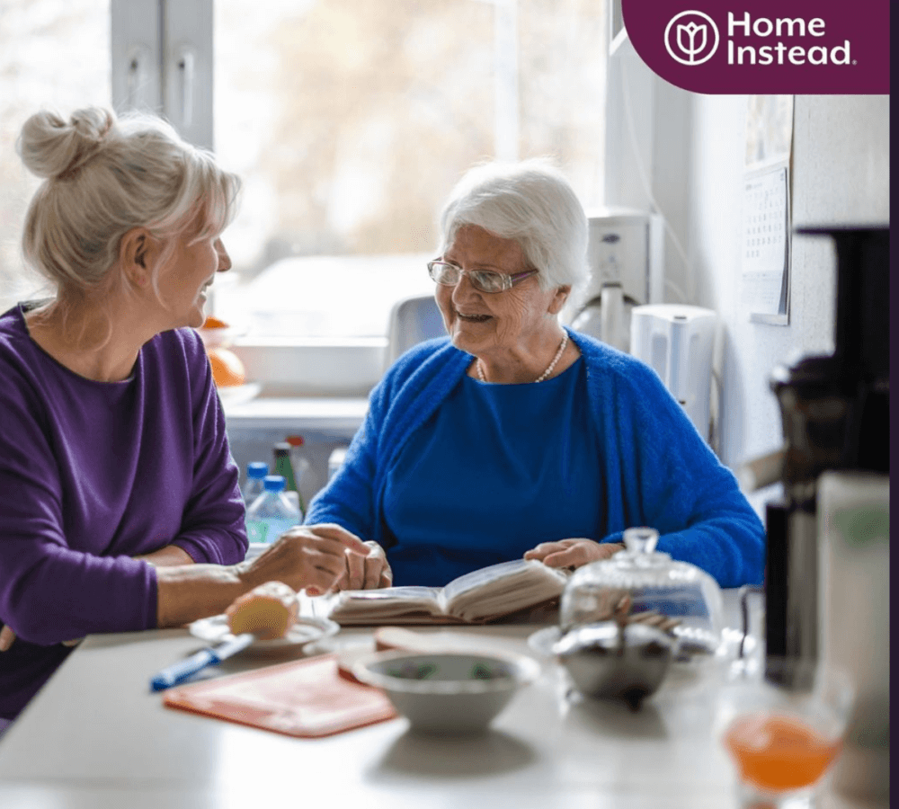 Two elderly women are sitting at a table, smiling and talking, with one holding a book. With Home Instead logo. - Home Instead