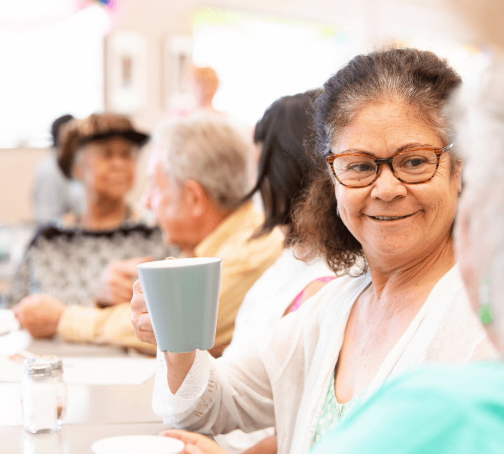 A woman with glasses smiles and holds a coffee cup while talking with other seniors in a bright, social setting. - Home Instead